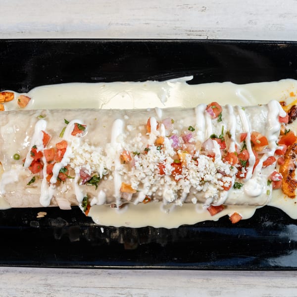 A large burrito covered in white sauce, pico de gallo, and crumbled cheese on a black plate.