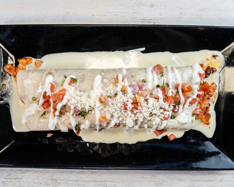 A large burrito covered in white sauce, pico de gallo, and crumbled cheese on a black plate.