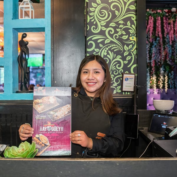 A smiling restaurant worker holds up a menu for "¡¡AY GÜEY!! Modern Mexican Cuisine" behind a counter.