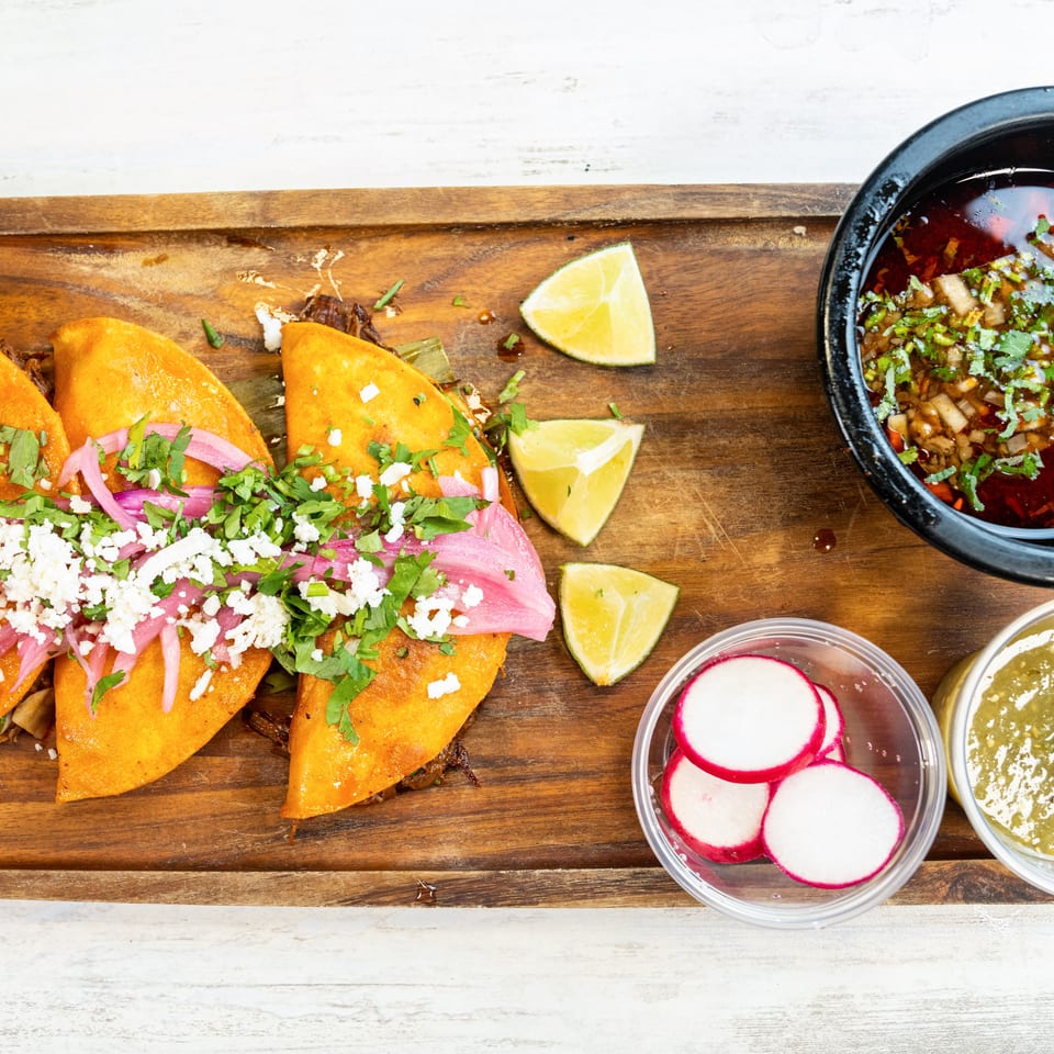 Three birria tacos with red onions, cilantro, and cotija cheese, with consommé, limes, radishes, and salsa.