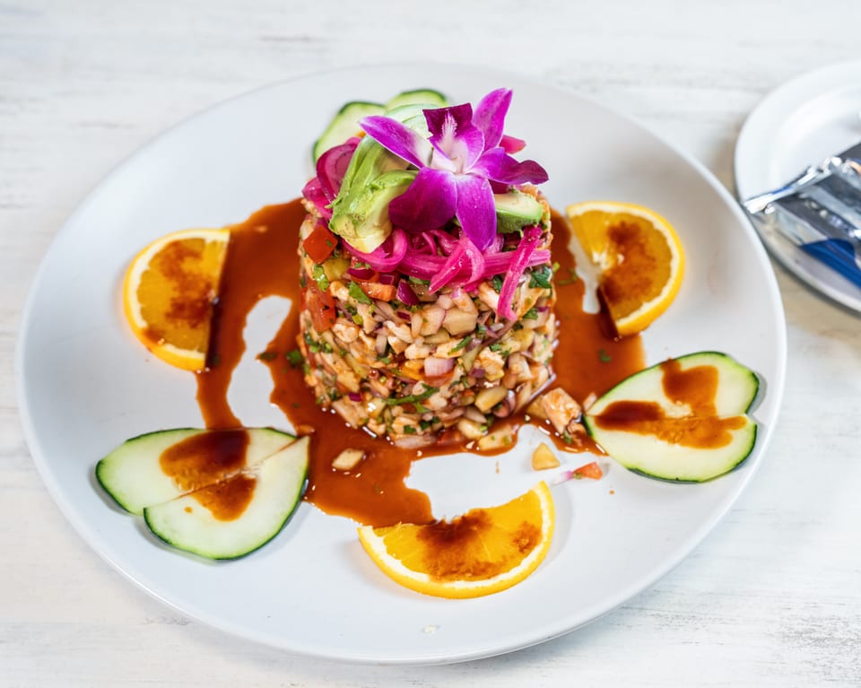 A vibrant ceviche tower with avocado, pickled onions, and an orchid, garnished with orange and cucumber slices.