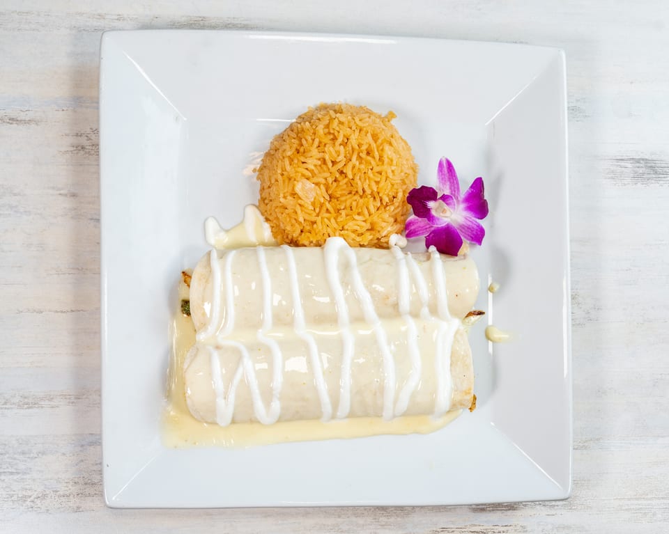Two enchiladas topped with white sauce, a side of rice, and a purple orchid garnish.