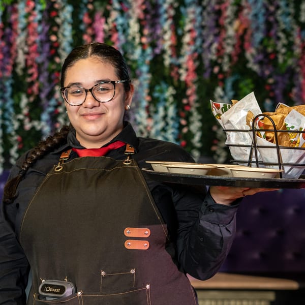 Smiling server in an apron holding a tray with tortilla chips and small bowls.