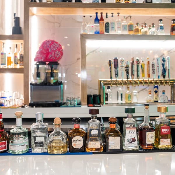 A bar counter displays a variety of tequila bottles, with beer taps and liquor shelves in the background.