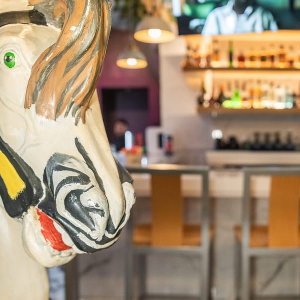 Painted carousel horse head with green eyes in foreground, blurred bar with bottles and stools in background.