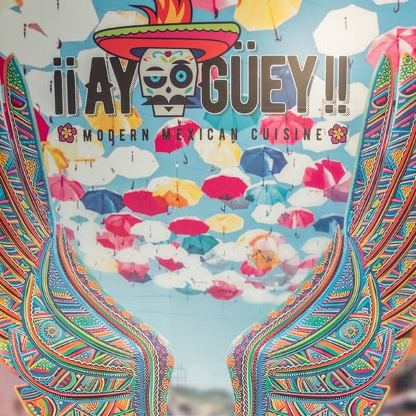 Colorful sign for "¡¡AY GÜEY!! Modern Mexican Cuisine" with a sugar skull logo and umbrella background.