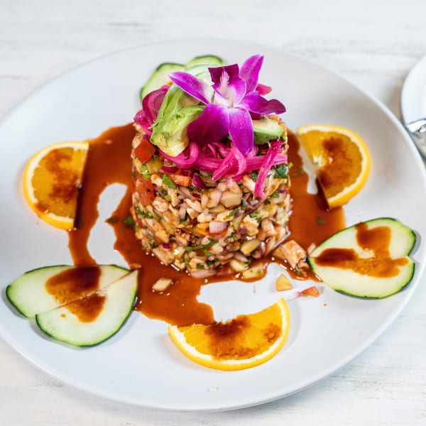 A vibrant ceviche tower garnished with avocado, pickled onions, and an orchid, surrounded by orange and cucumber slices.
