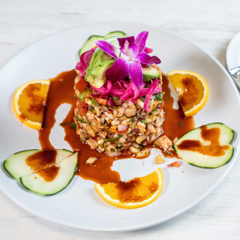 A vibrant ceviche tower garnished with avocado, pickled onions, and an orchid, surrounded by orange and cucumber slices.