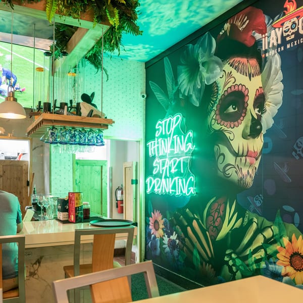 A man sits at a bar with a large TV showing soccer, and a neon sign on a mural.
