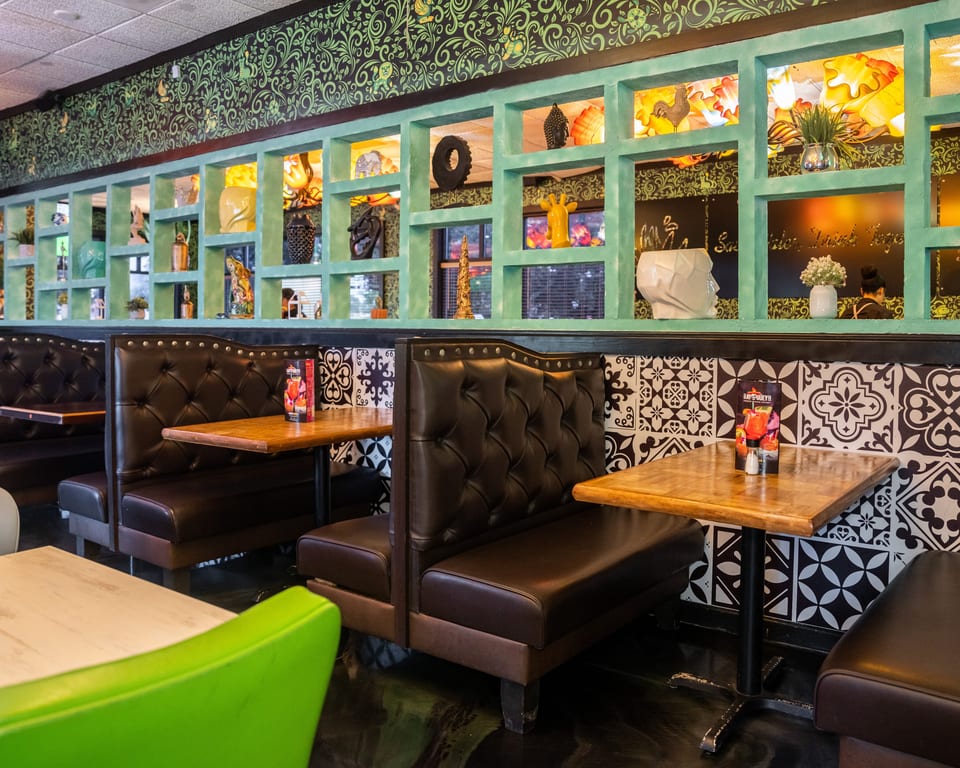 Restaurant interior with dark brown booths, wooden tables, and a decorative wall with green shelves and patterned wallpaper.