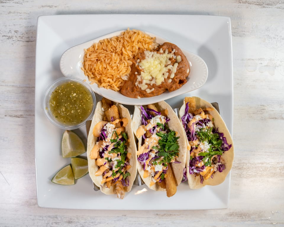 Three fish tacos with red cabbage, sauce, and cilantro, served with rice, beans, salsa, and lime wedges.