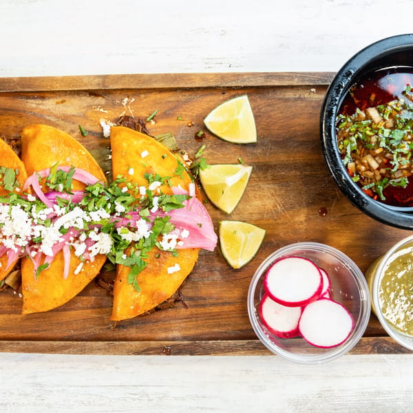 Three birria tacos with red onions, cilantro, and cotija cheese, with consommé, limes, radishes, and salsa.