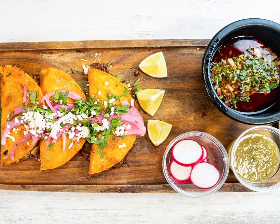 Three birria tacos with red onions, cilantro, and cotija cheese, with consommé, limes, radishes, and salsa.