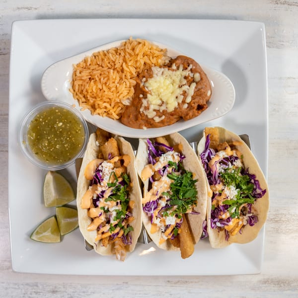 Three fish tacos with red cabbage, sauce, and cilantro, served with rice, beans, salsa, and lime.