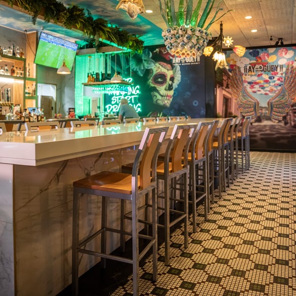 A vibrant bar interior with a long counter, stools, colorful murals, and neon signs.