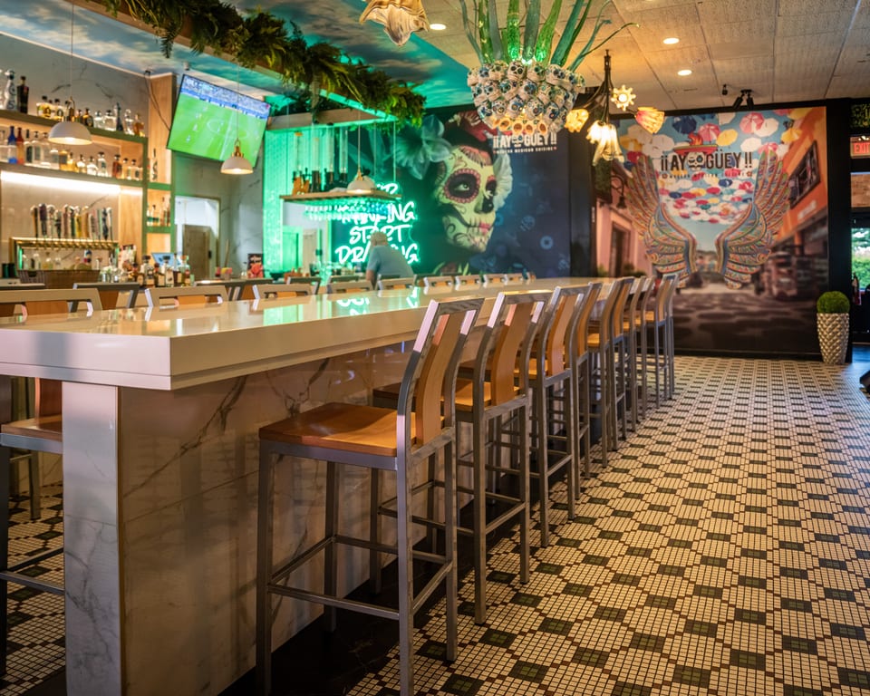 A vibrant bar interior with a long counter, stools, colorful murals, and neon signs.