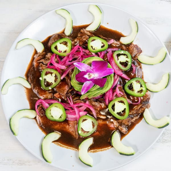 A dish with sliced meat in a dark sauce, topped with red onions, jalapeños, avocado, and a purple orchid.