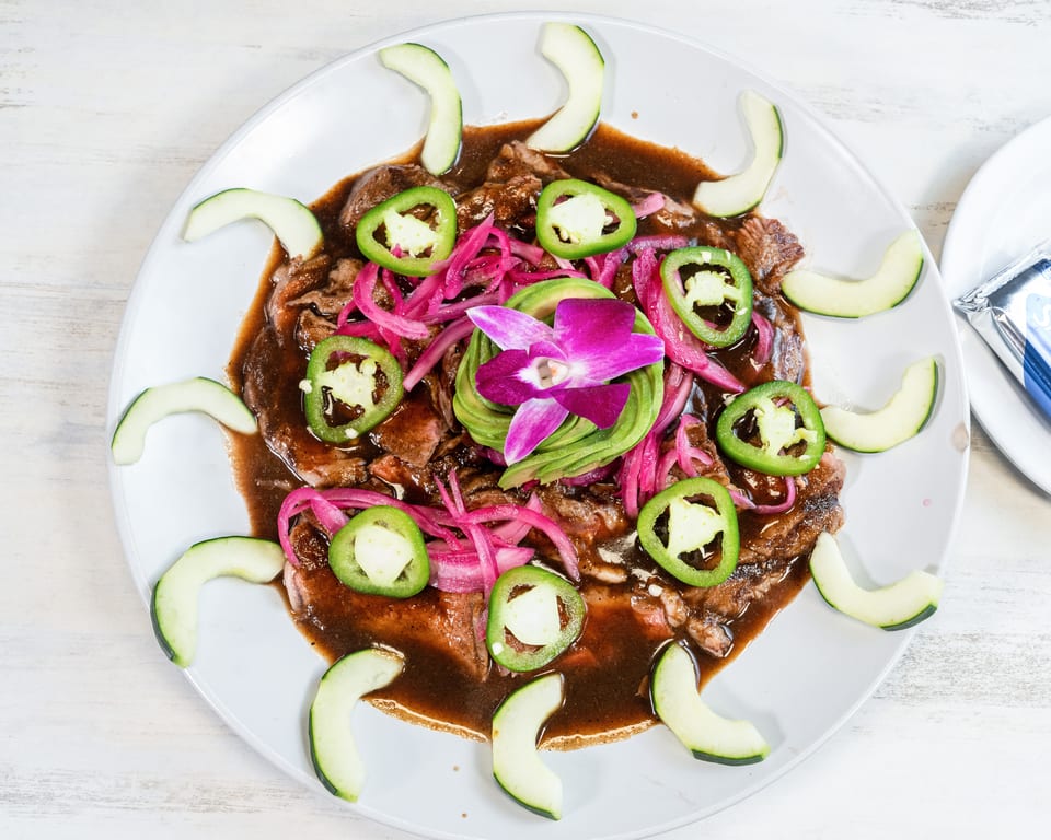 A dish with sliced meat in a dark sauce, topped with red onions, jalapeños, avocado, and a purple orchid.