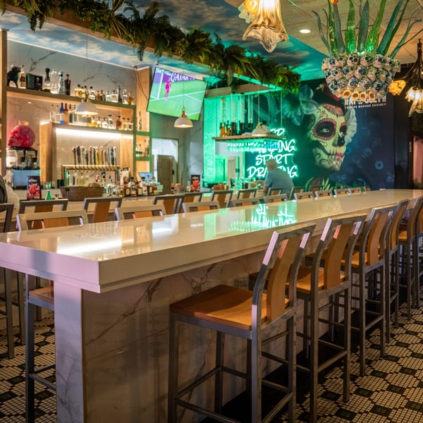 A vibrant bar interior with a long white counter, bar stools, and colorful decor including a skull mural.