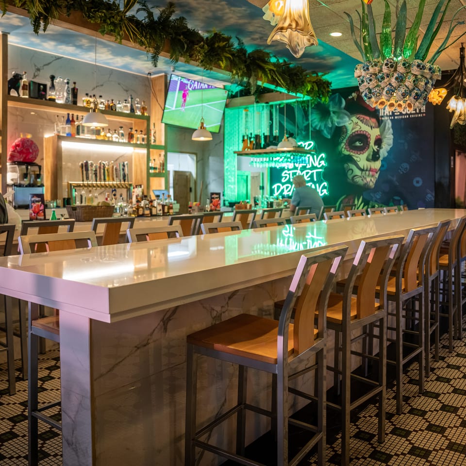 A vibrant bar interior with a long white counter, bar stools, and colorful decor including a skull mural.