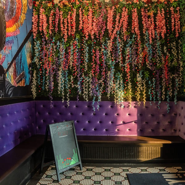 Restaurant interior with purple velvet booth seating, a floral wall, and a "Bienvenidos" chalkboard sign.