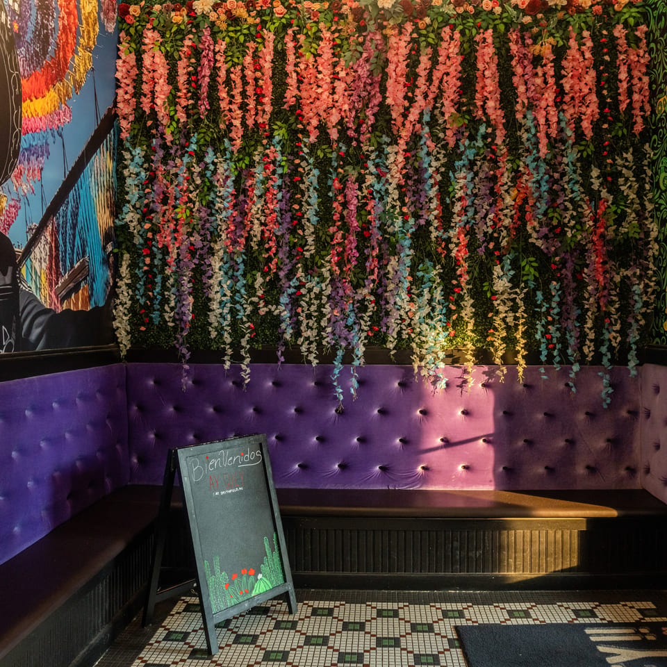 Restaurant interior with purple velvet booth seating, a floral wall, and a "Bienvenidos" chalkboard sign.