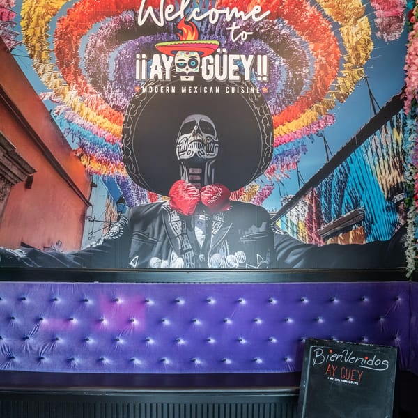 A vibrant mural of a mariachi skeleton and colorful banners above a purple tufted booth.