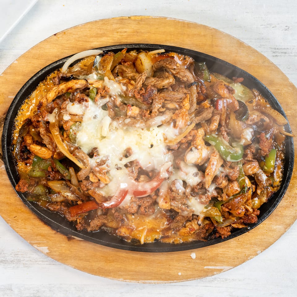 Sizzling fajitas with melted cheese, chicken, beef, peppers, and onions on a wooden platter.