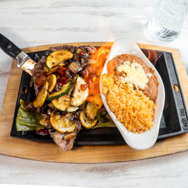 Sizzling steak fajitas with grilled vegetables, rice, and refried beans on a wooden board.