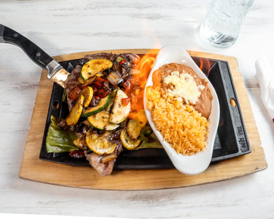 Sizzling steak fajitas with grilled vegetables, rice, and refried beans on a wooden board.